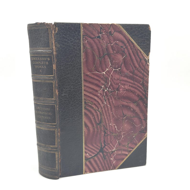 Puede incluir: Libro antiguo con lomo y cubierta de cuero negro, titulado "Emerson's Complete Works" y "Lectures Biographical Sketches". La cubierta del libro presenta un dise&ntilde;o veteado en burdeos y crema.