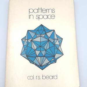 May include: A softcover book titled Patterns in Space by Col. R.S. Beard. The off-white cover features a large, central geometric illustration composed of interconnected blue and grey triangles forming a complex, multi-faceted star-like shape.