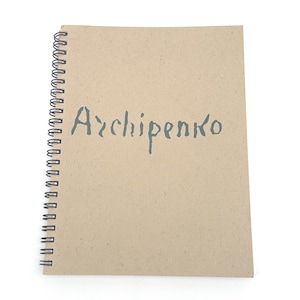 May include: A spiral-bound notebook with a kraft paper cover. The word "Archipenko" is stenciled in a muted teal color across the front. The notebook is open, displaying the cover and spiral binding.