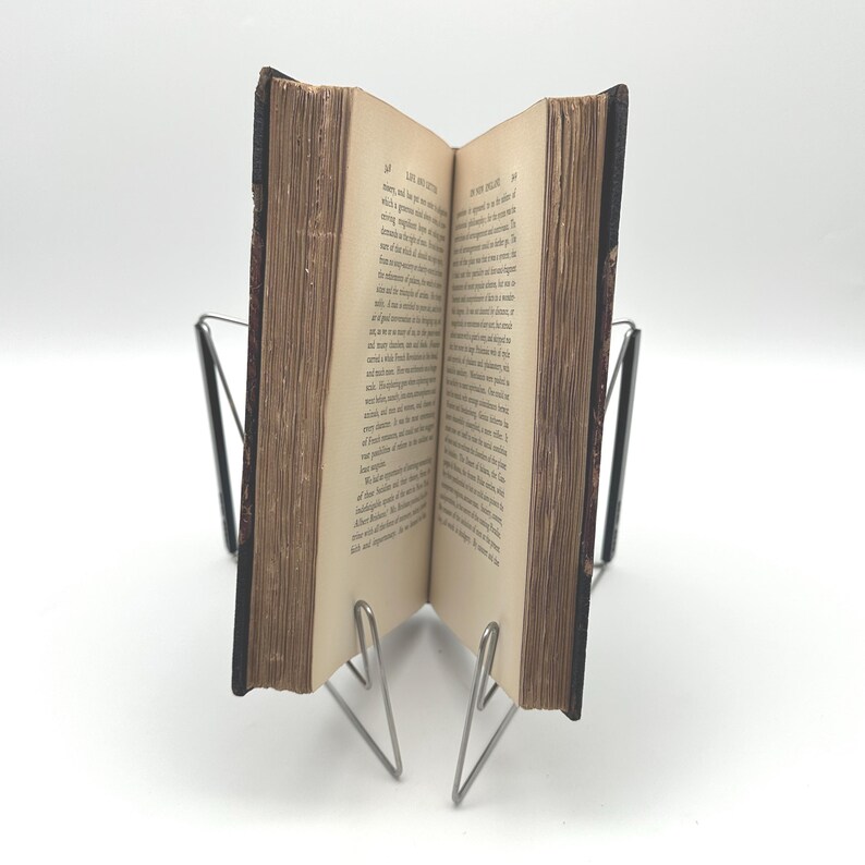 Puede incluir: Un libro antiguo abierto, con p&aacute;ginas amarillentas y texto visible, sostenido por un soporte de libro de metal plateado. El libro tiene una cubierta marr&oacute;n oscuro y las p&aacute;ginas est&aacute;n ligeramente curvadas.