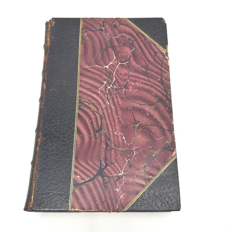 Puede incluir: Libro antiguo con lomo y esquinas de cuero negro. La cubierta presenta un patr&oacute;n veteado burdeos y crema con ribete dorado. Los bordes del libro muestran signos de desgaste, lo que sugiere su antig&uuml;edad y uso.