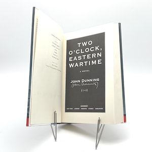 May include: An open book titled "TWO O'CLOCK, EASTERN WARTIME" by John Dunning. The book's cover is black with white text. The book is propped open by a silver metal book stand. The book's pages are off-white.