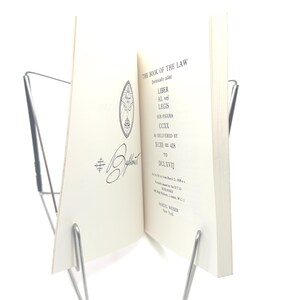 May include: An open book titled "THE BOOK OF THE LAW" in black ink, held by a silver metal book stand. The book's pages are off-white, with text and a decorative emblem. The book is displayed against a white background.