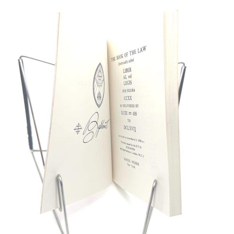 May include: An open book titled "THE BOOK OF THE LAW" in black ink, held by a silver metal book stand. The book's pages are off-white, with text and a decorative emblem. The book is displayed against a white background.