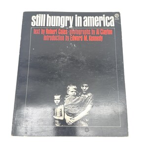 Puede incluir: Un libro titulado "still hungry in america" con texto de Robert Coles, fotografías de Al Clayton y una introducción de Edward M. Kennedy. La portada presenta una fotografía en blanco y negro de tres personas.