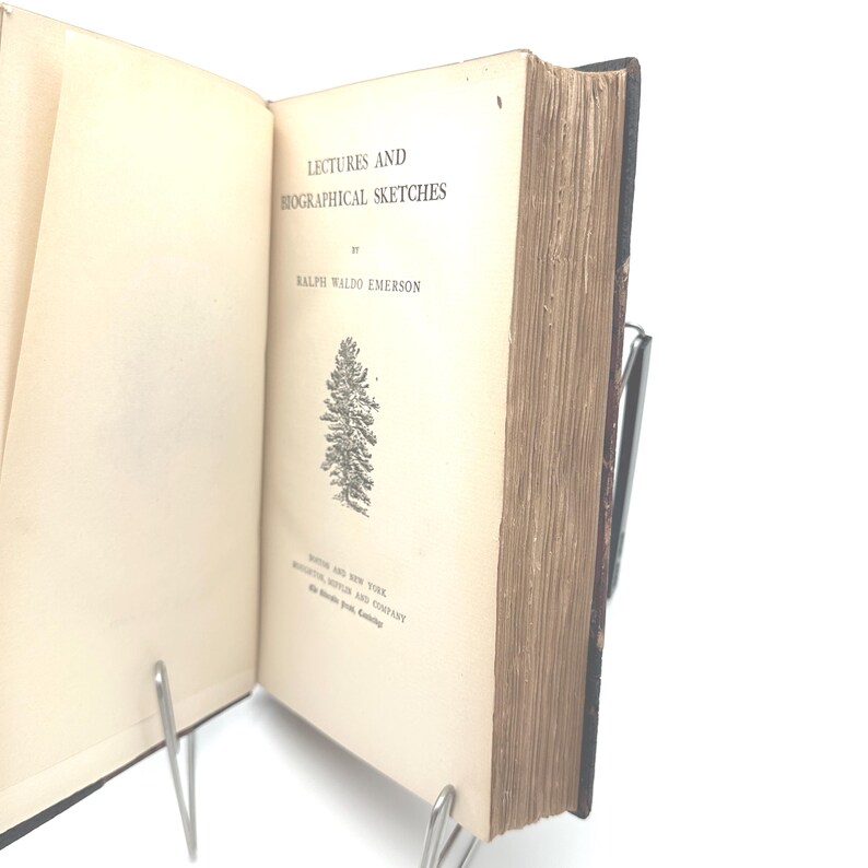 Puede incluir: Un libro abierto con el t&iacute;tulo "Lectures and Biographical Sketches" de Ralph Waldo Emerson. El libro tiene un aspecto vintage con p&aacute;ginas envejecidas de color blanquecino y una ilustraci&oacute;n detallada de un &aacute;rbol. El libro est&aacute; sostenido abierto por un atril met&aacute;lico.