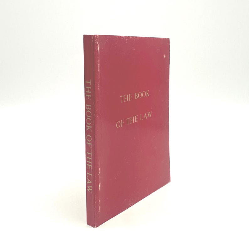 May include: A maroon-colored book titled "THE BOOK OF THE LAW" in gold lettering. The book's spine also features the title. The book is standing upright against a white background.