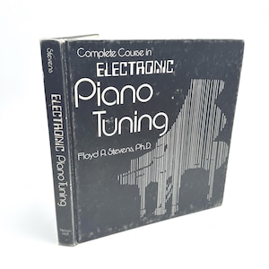 Op de afbeelding: Een vintage boek getiteld "Complete Course in Electronic Piano Tuning" door Floyd A. Stevens, Ph.D. De omslag is donkerblauw met witte tekst en een piano-illustratie. De rug leest "Electronic Piano Tuning".