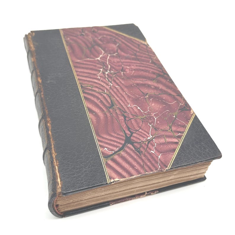 Puede incluir: Un libro antiguo con lomo y esquinas de cuero negro, y una cubierta veteada burdeos y crema. El libro tiene ribetes dorados y p&aacute;ginas amarillentas, lo que sugiere su edad y valor hist&oacute;rico.