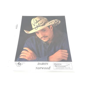 May include: A photograph of Daron Norwood wearing a cowboy hat with signatures. The image is a portrait of a man with long hair and a mustache, wearing a blue shirt. The text "Daron Norwood" is printed at the bottom.