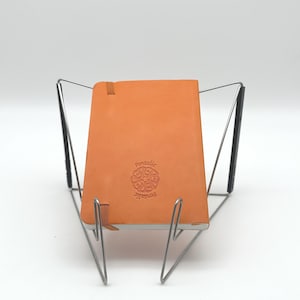 May include: An orange leather notebook with the word "Pentallic" embossed on the cover, resting on a silver metal stand. The notebook has a ribbon bookmark. The stand has a unique, geometric design.