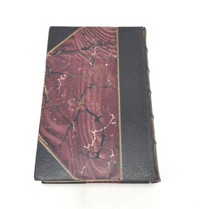 Puede incluir: Un libro antiguo de tapa dura con una cubierta veteada en burdeos y negro. La cubierta presenta un ribete dorado y un lomo negro. Las p&aacute;ginas del libro est&aacute;n encuadernadas y los bordes est&aacute;n ligeramente desgastados, lo que sugiere edad y uso.
