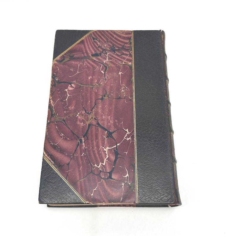 Puede incluir: Un libro antiguo de tapa dura con una cubierta veteada en burdeos y negro. La cubierta presenta un ribete dorado y un lomo negro. Las p&aacute;ginas del libro est&aacute;n encuadernadas y los bordes est&aacute;n ligeramente desgastados, lo que sugiere edad y uso.
