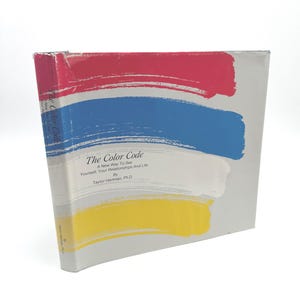 May include: A hardcover book titled "The Color Code" with a gray cover and colorful brushstroke design. The cover features red, blue, white, and yellow strokes. The book's title and author's name are printed on the front.