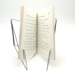 May include: An open book with handwritten text, propped up by a silver metal book stand. The pages are off-white, and the text is in black ink. The stand has a minimalist design, with thin, curved supports.