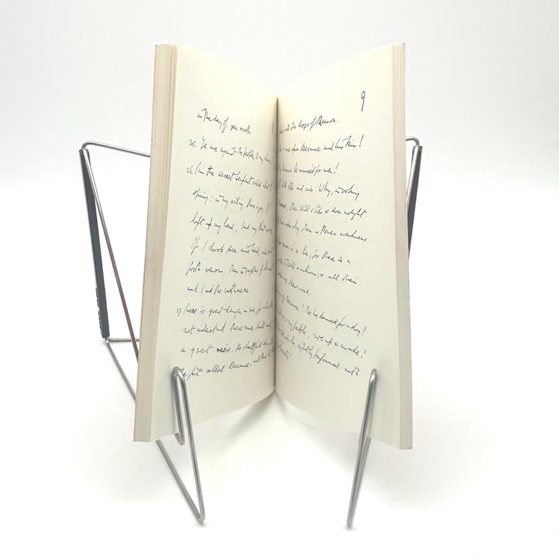May include: An open book with handwritten text, propped up by a silver metal book stand. The pages are off-white, and the text is in black ink. The stand has a minimalist design, with thin, curved supports.