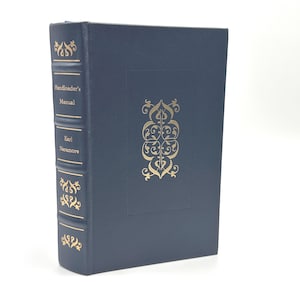 Puede incluir: Un libro de tapa dura azul marino titulado "Handloader's Manual" de Earl Naramore. El libro presenta detalles decorativos dorados y texto en el lomo y la portada, con un diseño ornamentado en el centro.