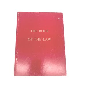 May include: A red book with the title "THE BOOK OF THE LAW" in white text. The book has a slightly worn appearance, with visible wear on the edges and corners. The cover is a solid, deep red color.