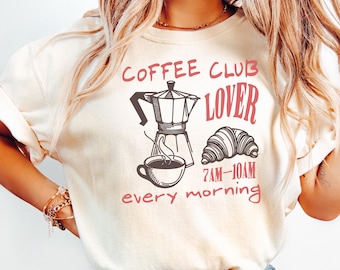 May include: Cream-colored t-shirt with a coffee-themed graphic. The design includes a coffee maker, a cup of coffee, and a croissant, with the words "Coffee Club Lover" and "7AM-10AM every morning" in red.