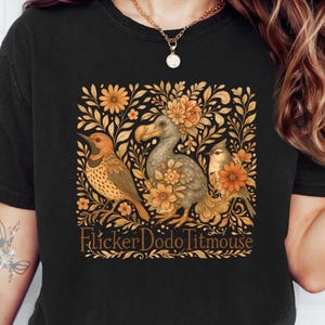 May include: Black t-shirt featuring a detailed illustration of three birds and floral elements in shades of orange and brown. The text "Flicker Dodo Titmouse" is below the illustration. The shirt is made of a soft material.