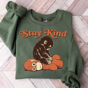 May include: An olive green sweatshirt with the words "Stay Kind" in orange script. The graphic features a brown monkey hugging an orange teddy bear. The sweatshirt is folded on a wooden surface.