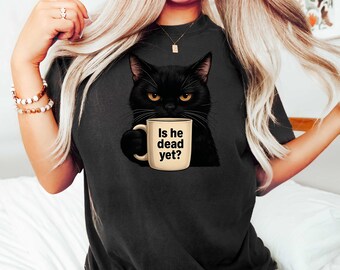 May include: Black t-shirt featuring a graphic of a black cat holding a mug. The mug has the text "Is he dead yet?" printed on it. The cat has yellow eyes and is centered on the shirt.