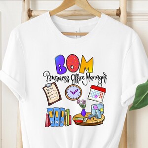 May include: White t-shirt with a colorful graphic design. The design includes the text "BOM Business Office Manager" with office-related illustrations such as a clock, calendar, and file folders. The shirt is hanging on a wooden hanger.