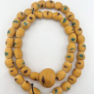 Puede incluir: Un collar hecho de cuentas amarillas-anaranjadas en forma de barril con detalles en marrón oscuro y verde azulado. El collar tiene una cuenta central grande y un cierre cilíndrico blanco. Las cuentas están ensartadas en un cordón negro.