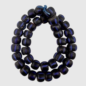 Puede incluir: Un collar circular de cuentas oscuras con acabado mate, cada una con una fina línea vertical de color azul vibrante que rodea el centro. Las cuentas están ensartadas en un cordón azul fino, formando un bucle continuo de formas redondeadas.