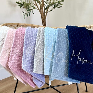 May include: Seven personalized baby blankets in various colors, including white, pink, lavender, light blue, and navy blue. Each blanket features a different name embroidered in white thread.