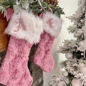 May include: Two pink fuzzy Christmas stockings with white faux fur cuffs. The stockings are decorated with pink glitter letters, one with the letter "A" and the other with the letter "E".