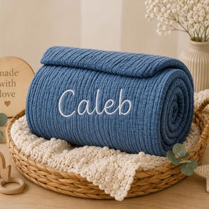 May include: A rolled blue muslin blanket with a textured, crinkled surface featuring the name Caleb embroidered in white script lettering. The blanket is placed inside a woven wicker basket lined with a cream-colored, bobbled knit fabric.