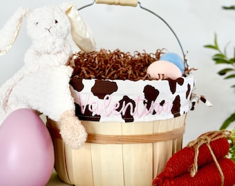May include: A white plush bunny with brown eyes sits in a brown and white cow print Easter basket with a pink egg, blue egg, and red carrots. The basket is labeled "Valentina".