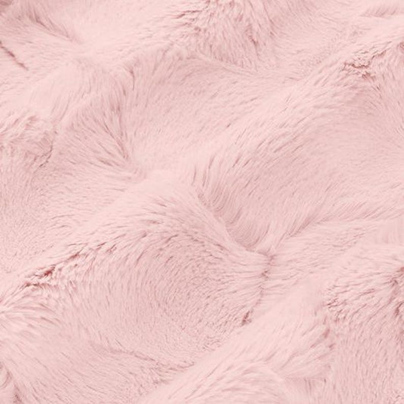 May include: A close-up shot of a soft, pink faux fur fabric. The plush material has a wavy texture with long, flowing fibres. The colour is a light, pastel pink, creating a cosy and inviting aesthetic. The fabric appears to be a blanket or throw.