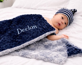 Może przedstawiać: Granatowo-szary kocyk dla dziecka z białym haftem z imieniem "Declan". Kocyk ma teksturowaną, pluszową powierzchnię. Widoczna jest również czapka w granatowo-białe paski.