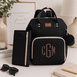 May include: A black backpack with gold-tone zippers and hardware, featuring a quilted front pocket with an embroidered monogram of the letters CGM. The set includes a matching rectangular zippered pouch and a small circular zippered accessory case.