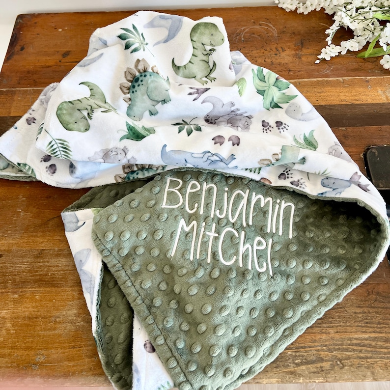 Op de afbeelding: Een wit en groene babydeken met een dinosaurusprint en de naam "Benjamin Mitchel" in het wit geborduurd. De deken heeft een groene, getextureerde onderkant.