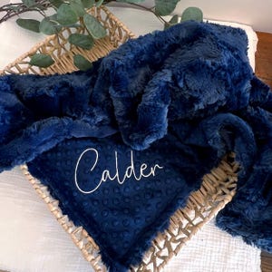 May include: A navy blue baby blanket with the name "Calder" embroidered in white script. The blanket has a textured, plush surface and a dotted pattern on the bottom layer. It is displayed in a woven basket.