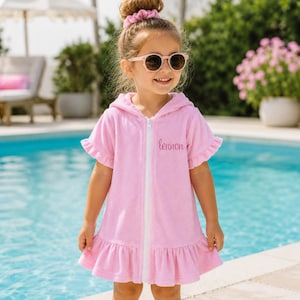 Peut inclure: Une robe de bain rose à capuche avec une fermeture éclair blanche et une bordure à volants. La robe a des manches courtes et le nom "lennon" brodé sur le devant. L'enfant porte des lunettes de soleil et un chouchou rose assorti.