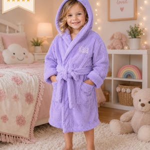 May include: A plush lavender hooded bathrobe featuring long sleeves, two front pockets, and a matching tie belt. The left chest area is embroidered with the name Kaia in a white script font. The robe is made of a soft, textured fabric.