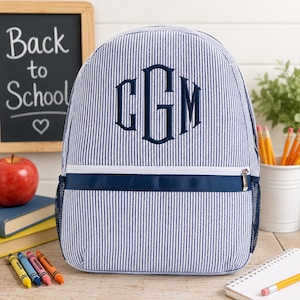 May include: Blue and white striped backpack with a navy blue monogram and trim. The backpack has a zippered front pocket and mesh side pocket. A chalkboard with "Back to School" is in the background.