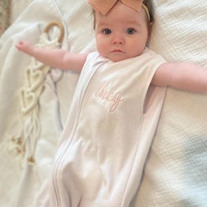 May include: A baby wearing a white sleep sack with the name "Lucy" embroidered in pink. The baby has a peach-colored bow headband. A wooden teething ring and macrame heart decor are in the background.