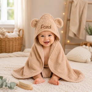 May include: A light brown hooded baby towel made of soft terry cloth fabric. The hood features two small rounded bear ears and the embroidered name Ford in dark brown thread. The towel is draped over a baby, covering the shoulders and torso.