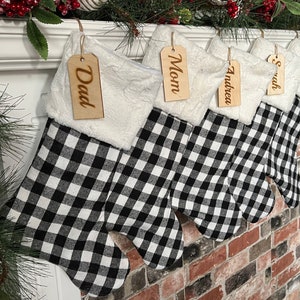 May include: Five black and white buffalo plaid Christmas stockings with white faux fur trim and wooden name tags. The tags read "Dad", "Mom", "Andrea", "Sarah", and "Broox".