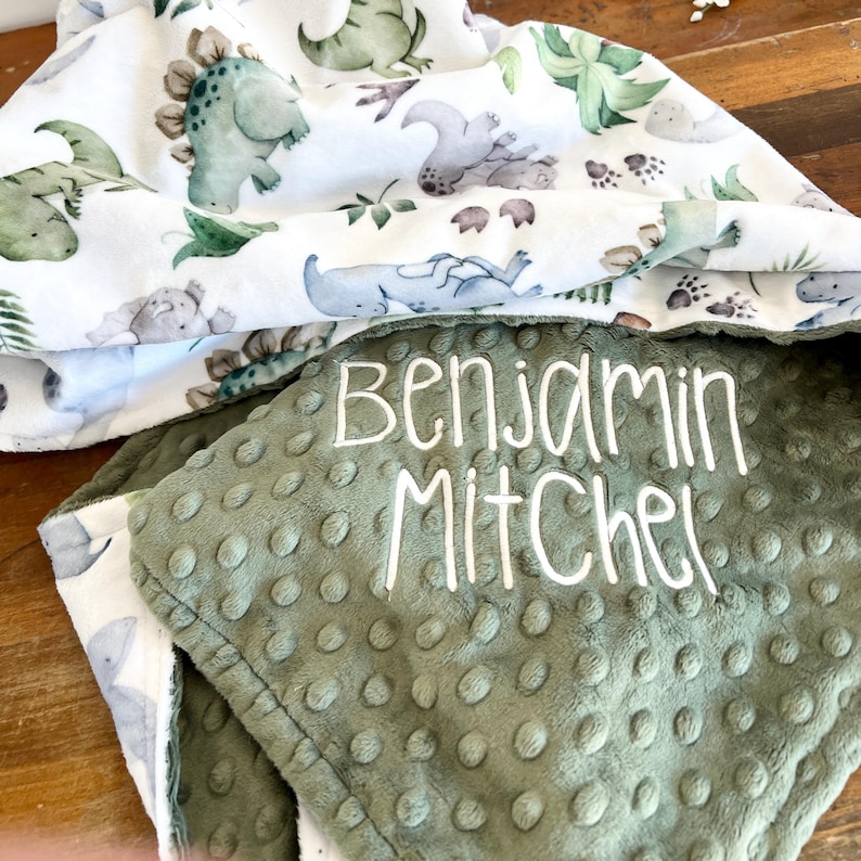 Op de afbeelding: Een babydeken met een witte dinosaurusprint en een groene, getextureerde onderkant. De naam "Benjamin Mitchel" is in het wit op de groene stof geborduurd. De deken is opgevouwen en toont beide zijden.