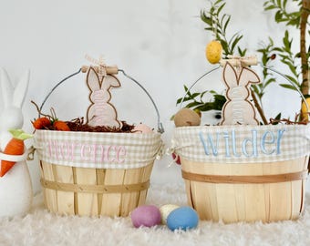 May include: Two round wooden baskets with metal handles and beige gingham fabric liners. One basket features the name Florence in pink embroidery, the other Wilder in blue. Each has a wooden bunny tag and decorative filler.