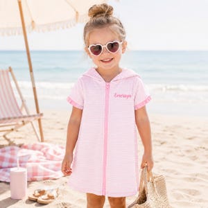 May include: A young child wearing a pink and white striped hooded beach cover-up with a zipper. The cover-up has short sleeves and the name "Emersyn" embroidered on the chest. The child is wearing heart-shaped sunglasses and holding a woven tote bag on a sandy beach.