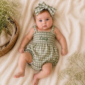 May include: A baby wearing a green and white gingham romper and matching headband with a bow. The romper has the name "Lucy" embroidered on the chest. The baby is lying on a cream-colored blanket.
