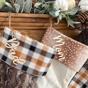 May include: Two Christmas stockings with personalized wooden tags. The stocking on the left is brown faux fur with a black and tan plaid cuff. The stocking on the right is white faux fur with a brown and white spotted cuff. The tags say "Dad" and "Mom".