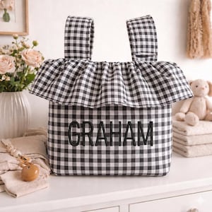 May include: A black and white gingham fabric tote bag with the name "GRAHAM" embroidered in black. The bag has two handles and a ruffled top. The bag is sitting on a white surface.